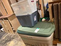 Stacked storage bins showing a large green Rubbermaid bin on bottom, a tan Rubbermaid bin in the middle, a gray Rubbermaid bin and clear plastic bin on top.