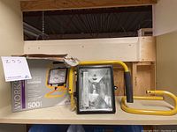 Globe Workshop 500 watt halogen work light with yellow metal stand and original box, showing the front of the work light fixture and packaging.