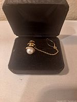 Tie tack in black velvet presentation box, showing pearl front, gold base, and safety chain.