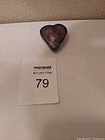 Top and front view of a purple handmade heart-shaped art glass piece placed on a white surface next to a lot number card