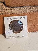 Front side of the RCMP Centennial quarter showing the mounted RCMP officer design encased in a white 2x2 coin holder with handwritten label.