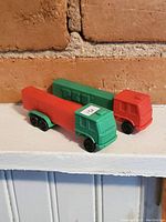 Photo showing two vintage toy trucks, one red and one green, positioned side by side on a white shelf against a brick wall.