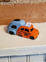 Two miniature Hot Wheels cars, orange and light blue, shown from side angles, placed on a white surface against a brick background.