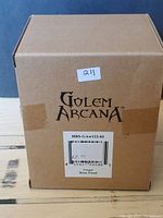 Photo of unopened cardboard box labeled Golem Arcana with sticker 211, showing product code HBS-GAw122-01 and name Urugal Bone Fiend.