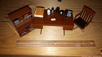 Miniature Marvel Spider-Man themed office desk with telephone, books, and small bottles, a brown office chair on wheels, and a brown bookcase with miniature books all on a wooden surface with measuring ruler.