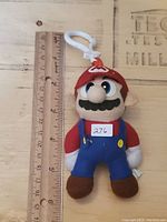 Front view of the Super Mario plush key chain next to ruler showing approx. 14 cm height. Features red hat with 'M', blue overalls, brown shoes, and plastic clip on top.