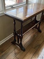 Full side view of the antique accent table showing spindle legs, carved decorative trim on the tabletop edge, and the carved support shelf under the tabletop.