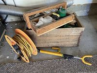 Wooden storage box with various items including a yellow extension cord reel, wooden toolbox, and long tool with yellow handle visible.