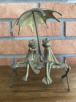 Front view of brass sculpture showing two frogs smiling, sitting on rustic bench holding umbrella.