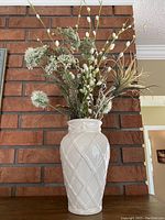 Front view of the tall white ceramic vase with glossy diamond lattice pattern and floral arrangement on top, set against a brick wall.
