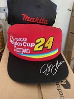 Close-up showing red NASCAR Winston Cup Champion 24 baseball hat with signature on brim, and a black Makita baseball cap behind it.