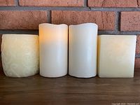 Four LED candles arranged side by side against a brick wall background showing their shapes, colors, and texture. One round candle has an embossed floral design, two smooth cylindrical candles show realistic melted tops with LED light on in one, and one square candle with slight embossing is present.