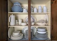 Three shelves inside wooden cupboard showing white china dinnerware including stacked plates, bowls, cups, coffee pot, serving dishes, and blue tea cozy