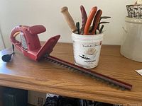 Red Homelite electric hedge trimmer lying on a surface next to a white plastic bucket filled with assorted hand gardening tools.