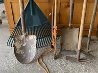 Full view of all gardening tools showing spade shovels, leaf rakes, tree trimmer secured with rope, saw, hand fork, and hoe.