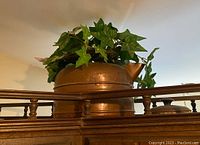 Copper kettle used as a planter with faux ivy plant spilling out, shown sitting on top of a wooden surface.