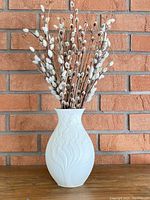 Full view of white Kaiser West Germany vase filled with dried pussy willow branches set against a brick wall background.