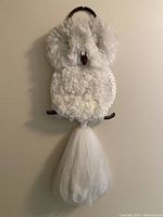 Full view of white fluffy macrame owl wall hanging mounted on a wall, showing the fluffy yarn body, wooden branch perch, and tassel tail.