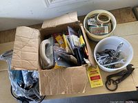 Photo of a cardboard box containing assorted tools and paint-related accessories, along with plastic buckets holding tape rolls and metal tools.
