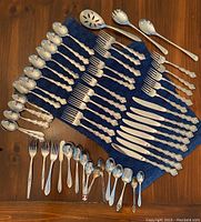 Photo of entire set of Oneida silverware laid out on blue cloth on wooden table showing multiple spoons, forks, knives, and serving utensils with ornate handles.