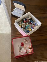 Photo showing three boxes of jewelry: one with clear rhinestone stud earrings in a small box, another white box filled with mixed earrings and pins in multiple colors and styles, and a red box with more colorful jewelry pieces including some pearl-like beads and enamel pins.