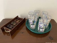 Wooden beverage carrier with eight colored shot glasses inside next to a green round tray with twelve floral plastic cups.