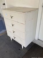 Front angled view of the vintage white dresser showing four drawers with metal pulls and tapered legs, two with casters.