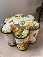 Top and angled front side view of a flower-shaped footstool showing the floral upholstery pattern in yellow, green, and brown on cream fabric and brown trim.