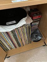 Wooden cabinet containing stacked vintage vinyl records, cassette tapes stacked upright, and some 45 RPM records stacked flat.
