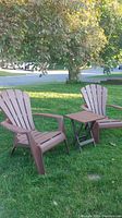 Two brown Adirondack style plastic chairs and one small brown folding side table on grass outdoors under a tree.