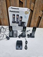 Three Panasonic cordless phones with charging bases and power adapters, displayed on a table with the box behind them. Shows the actual items included in the lot.