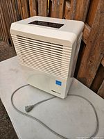 Front and side view of beige Uber-Haus dehumidifier with power cord coiled on ground beside it, showing ventilation grille and brand label.