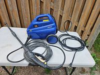 Side view of blue Simoniz electric pressure washer unit with power cord, coiled hose, and spray gun laid out on white table.