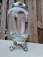 Glass beverage dispenser on decorative chrome stand with spigot and lid attached, placed outdoors.