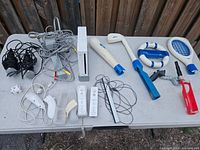 Overview of the entire lot showing Nintendo Wii console, controllers, cables, and Nerf branded accessories including tennis racquet, golf club, fishing rod, baseball bat, steering wheel, and microphones on white table.