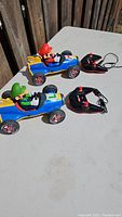 Two remote control cars featuring Mario and Luigi characters, each with its own wireless remote and charging cable, shown on table outdoors with sunlight.