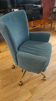 Side-angle view of the blue upholstered office chair showcasing the padded armrest and brass base with caster wheels on wooden floor.