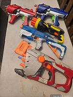 Five Nerf blasters displayed on table, two designed for round discs and three for darts, along with orange dart magazines.
