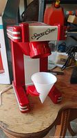 Front view of the red and white Nostalgia snow cone maker machine with a white reusable cone holder attached on a wooden surface.