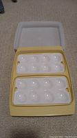 Yellow vintage Tupperware deviled egg holder with two white trays laid out holding indentations for 12 deviled egg halves total, placed on carpeted floor