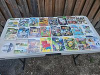 24 Nintendo Wii game cases displayed to show titles and cover art on a white outdoor table against wooden fence background.