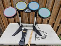 Xbox 360 Rockband drum kit featuring four multi-colored drum pads, control panel, pedal, and two drumsticks.