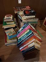 Pile of assorted hardcover and softcover books stacked on the floor, showing a mix of genres and titles.