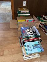 Photo shows assorted books in stacks and boxes on the floor including hard and soft covers, some titles visible.