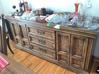 Full view of the oak buffet showing three drawers and two carved doors with storage inside, surface has various glassware items not included.