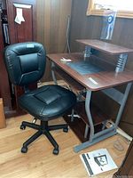 Desk and chair in a room with wood panel walls and wood laminate flooring. The desk has a wood top, a monitor shelf above, and a metal base with a side pull-out tray and lower shelf for storage. The chair is black with cushioned seat and back, mounted on casters, adjustable.