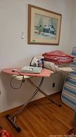 Metal folding ironing board with pink floral padded cover and a Black & Decker iron sitting on it. Several folded blankets and duvet nearby.