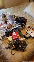 Various office supplies including pens, pencils, a digital thermostat, envelopes, file holders, batteries, chargers, binoculars, a flip phone, and glue on a bedspread.