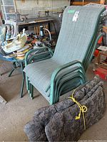 Photo showing four stackable green metal framed patio chairs with fabric mesh seats and backs, stacked together, alongside four dark floral patterned seat cushions tied with yellow rope on the ground.