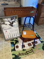 Overview photo showing woven plant stand, wooden frame calendar leaning against it, metal base chair with wooden seat nearby, and a small patterned floor mat on the floor.